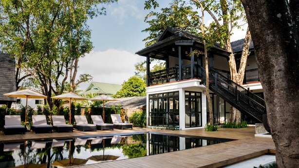 A pool area with lounge chairs, a wooden deck, and a building with a balcony and staircase. - MORA Boutique Hotel (Chiang Rai)