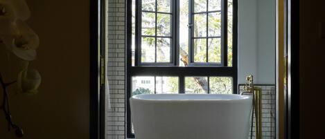 A bathroom with a freestanding bathtub, a window with blinds, and a tiled floor.