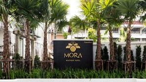 A sign for the MORA Boutique Hotel surrounded by palm trees and greenery. - MORA Boutique Hotel (Chiang Rai)