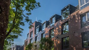 Ein modernes Gebäude mit mehreren Stockwerken, großen Fenstern und einem gepflegten Gartenbereich mit Bäumen und einem Weg.