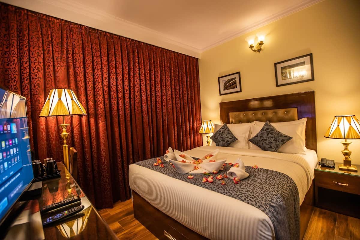 Quarto de hotel com uma cama, duas luminárias de cabeceira, uma televisão, uma secretária com um controle remoto e uma cortina.