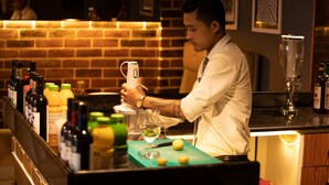 A bar area with a bartender preparing a drink, a brick wall, and various bottles and glasses on the counter. - Hotel Paramos Inn (Bengaluru)
