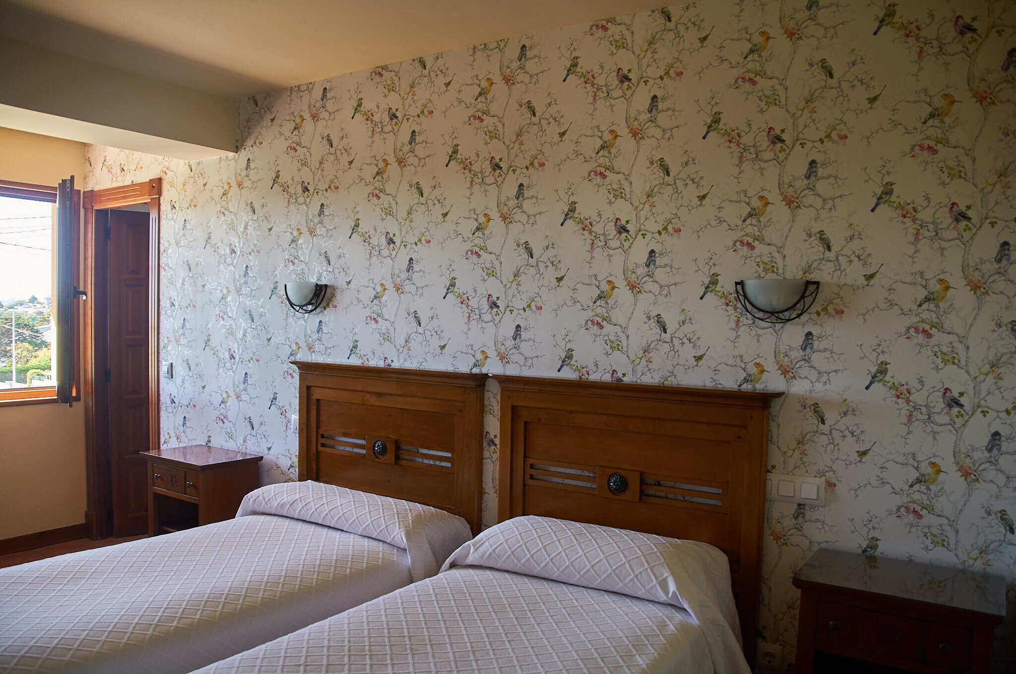 A bedroom with two beds, floral wallpaper, wooden headboards, bedside tables, and a window with a view.
