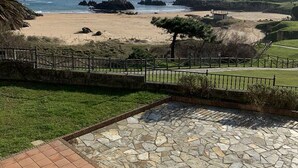 Eine Terrasse mit Steinbelag, Blick auf einen Strand mit Felsen und einem Zaun, sowie eine Rasenfläche.