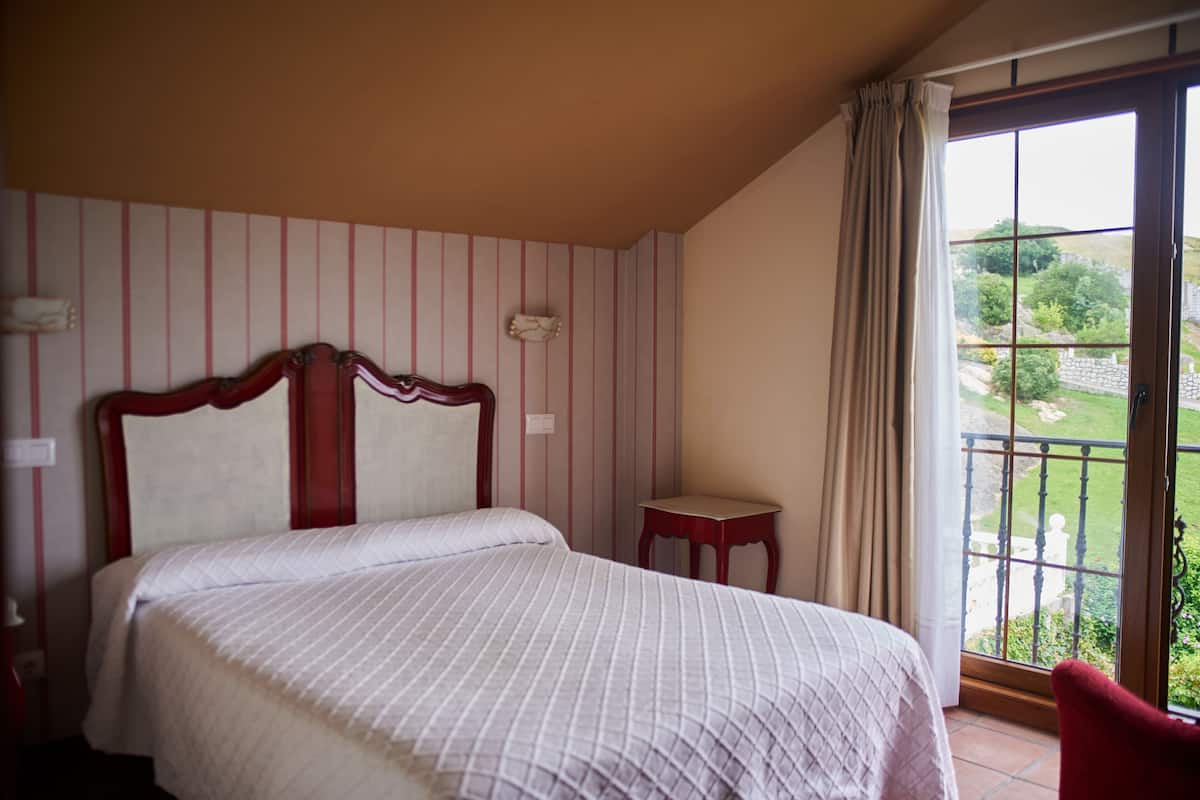 Une chambre à coucher comprenant un lit, une table de chevet, une petite table, une fenêtre donnant sur un paysage verdoyant et un rideau.