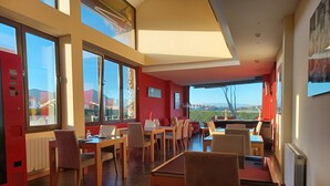 A modern dining area with multiple tables and chairs, large windows offering a view of the outdoors, and a red accent wall. - La Venta Del Mar (Pielagos)
