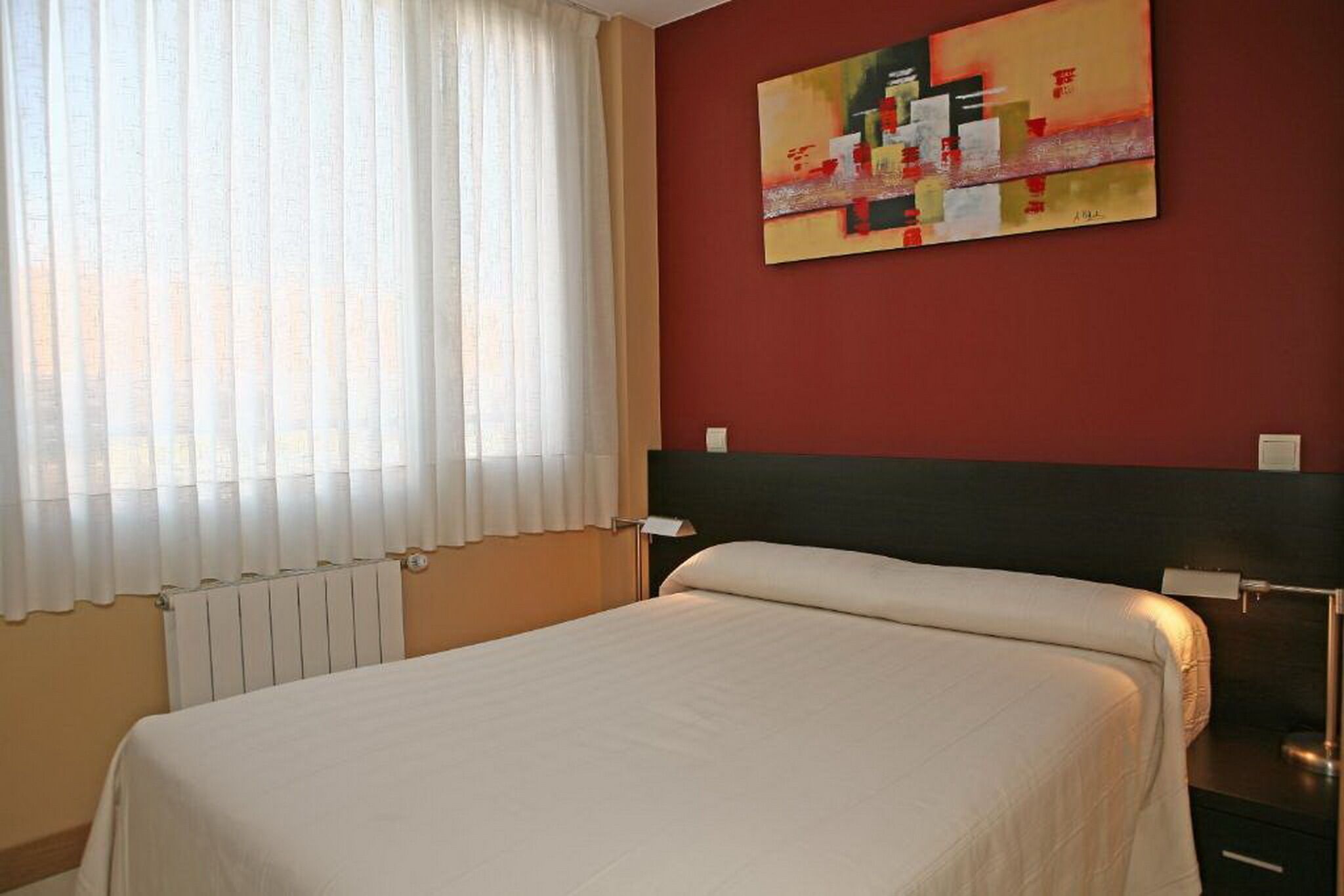 A hotel room with a bed, a radiator, a window with curtains, and a painting on the wall.
