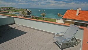 Un balcon avec une seule chaise en métal, un sol carrelé et une vue sur la mer et les bâtiments éloignés.