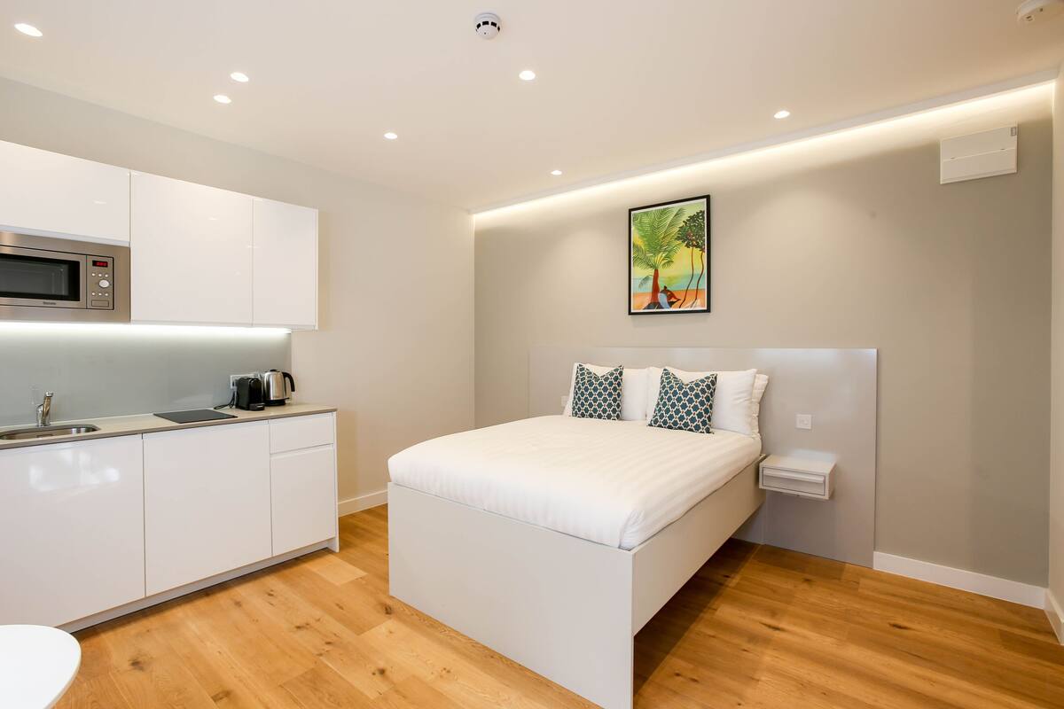 A modern bedroom with a bed, a bedside table, a nightstand, a wall-mounted air conditioner, and a kitchen area with a microwave.