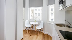 A modern kitchen with white cabinetry, a built-in oven, and a stainless steel sink. There is a small round table with two white chairs and a potted plant. - StayPrime Kensington Serviced Apartments (London)