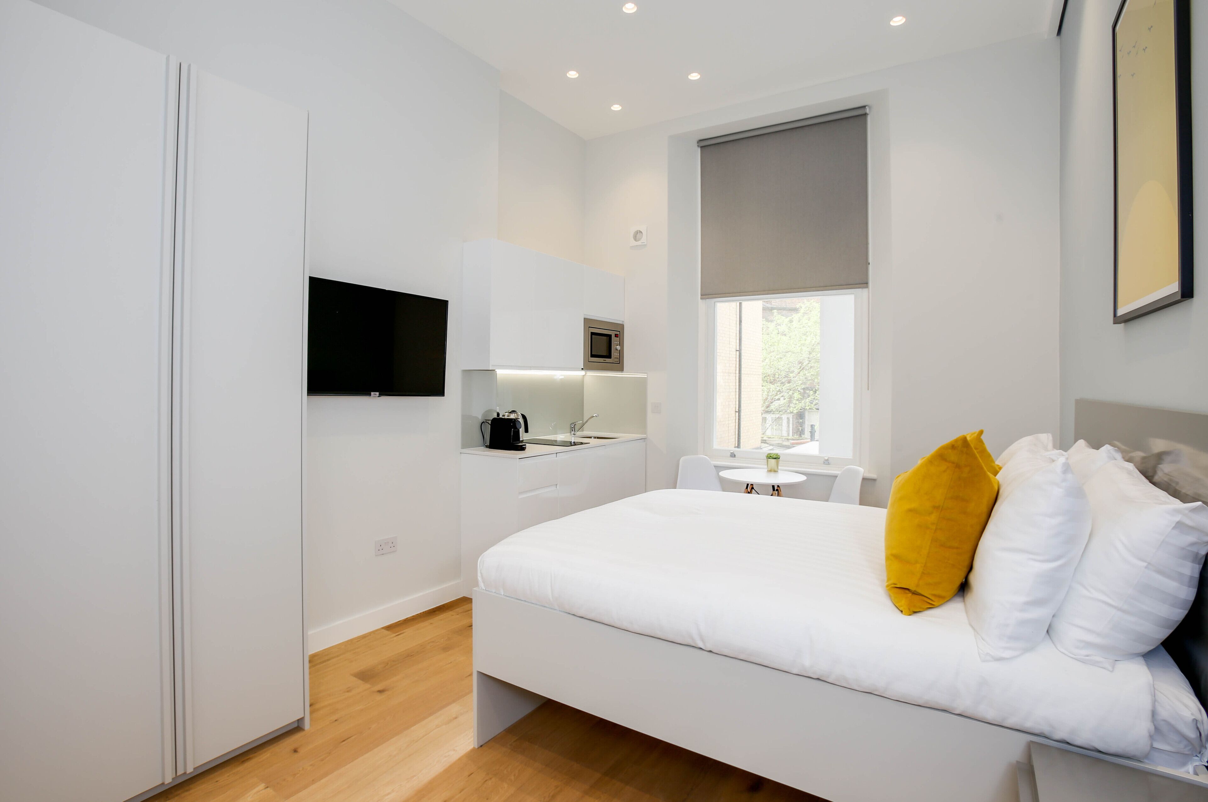 A modern bedroom with a bed, a TV mounted on the wall, a small kitchenette, and a window with a view.