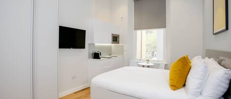 A modern bedroom with a bed, a TV mounted on the wall, a small kitchenette, and a window with a view.