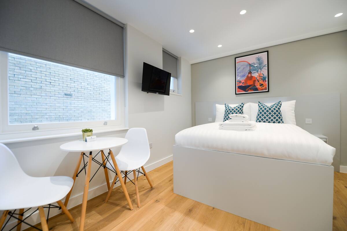 A modern hotel room with a bed, a TV, a small table with chairs, and a window with a view of a brick wall.
