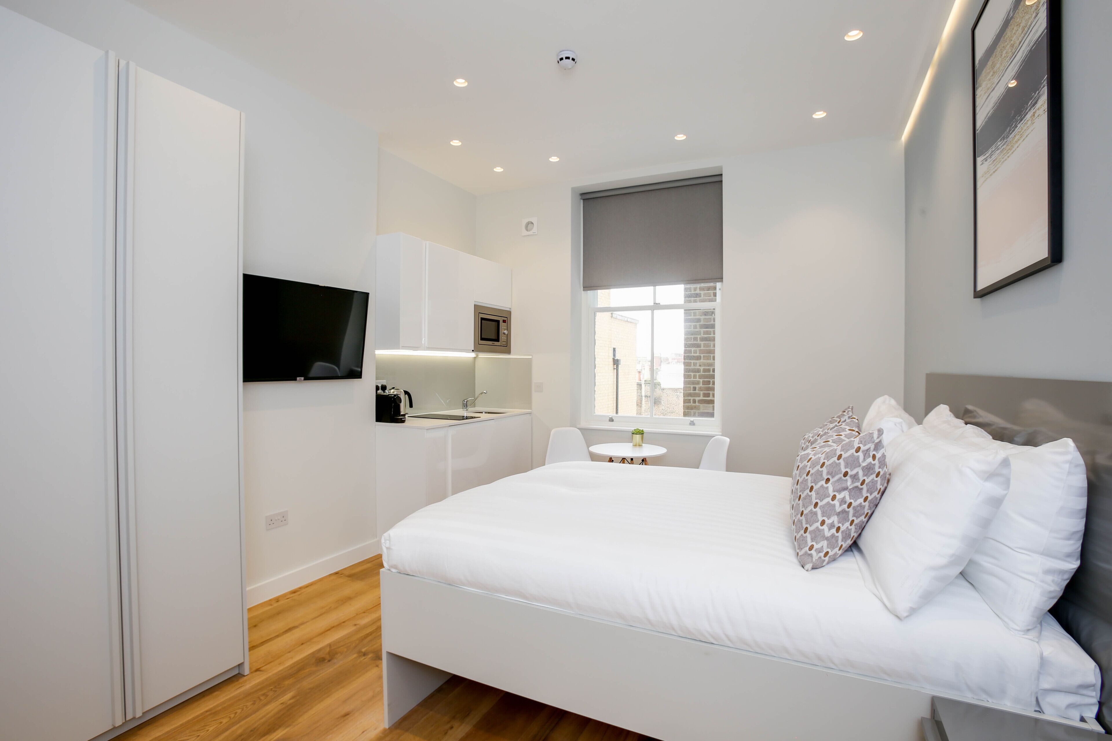 A modern bedroom with a bed, white walls, a small kitchen area, and a window.