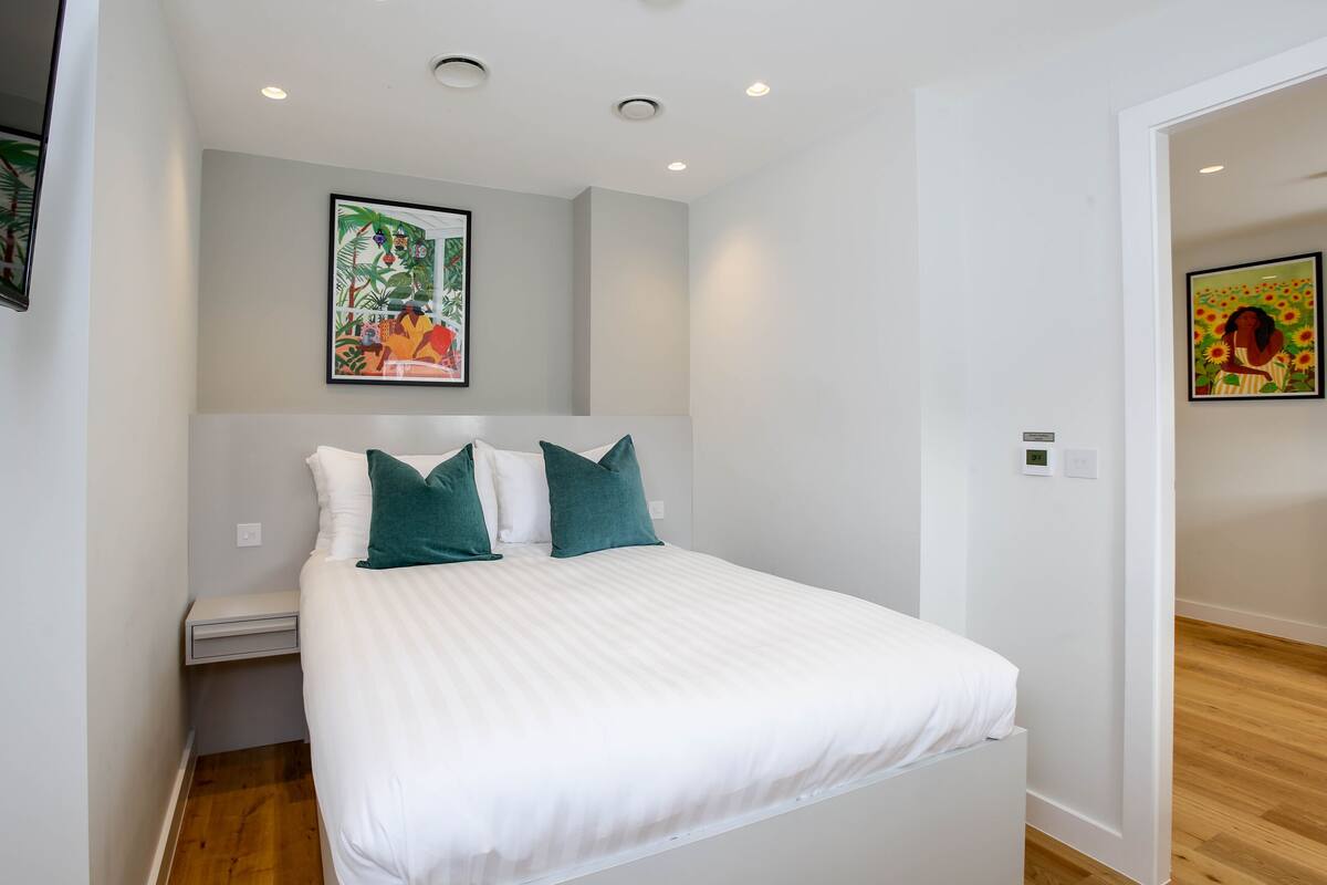 A neatly made bed with white bedding and teal pillows, a small bedside table, and a wall-mounted painting above it.
