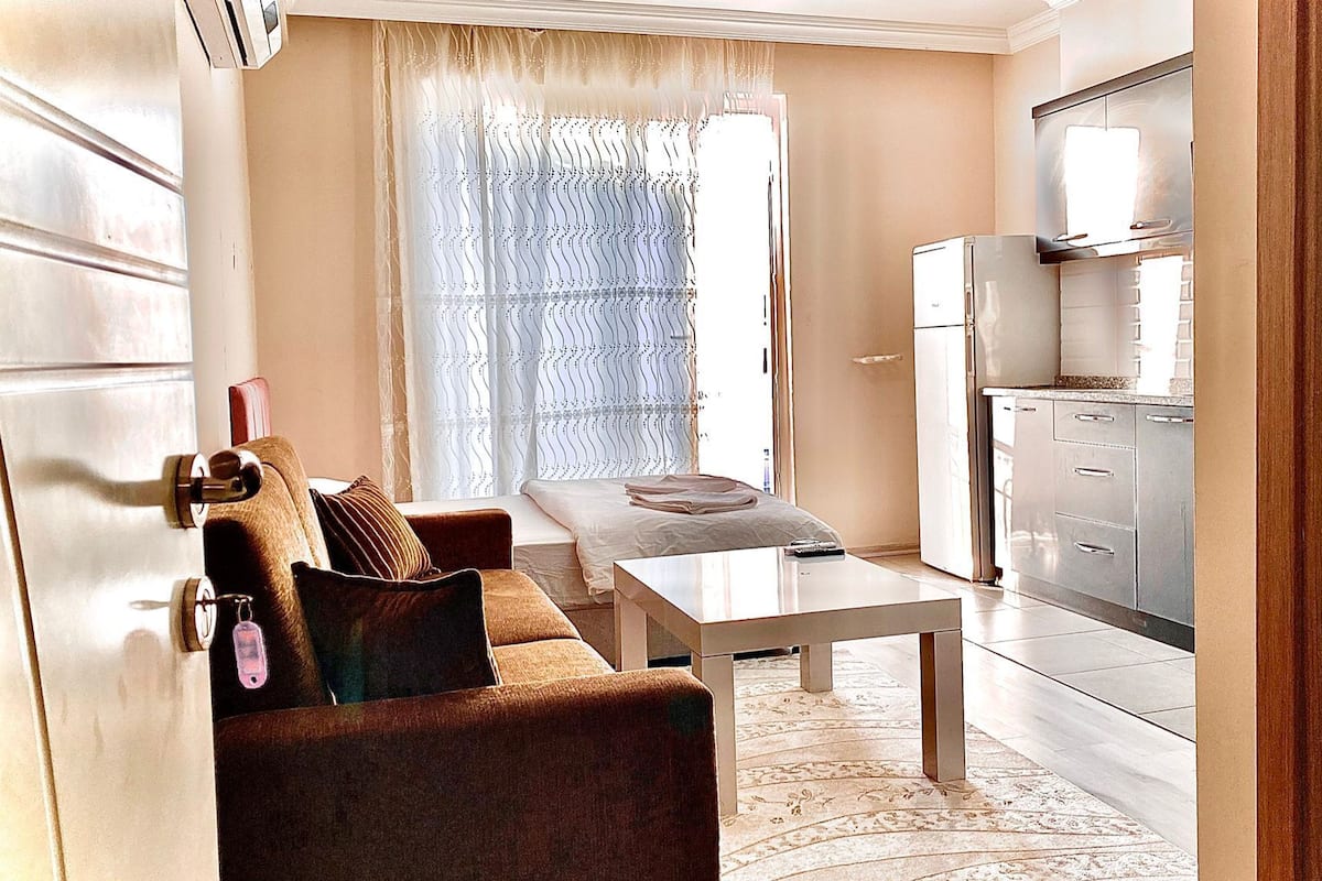 Um espaço compacto para viver, com sofá, mesinha, cozinha equipada com frigorífico e janela com cortinas estampadas.