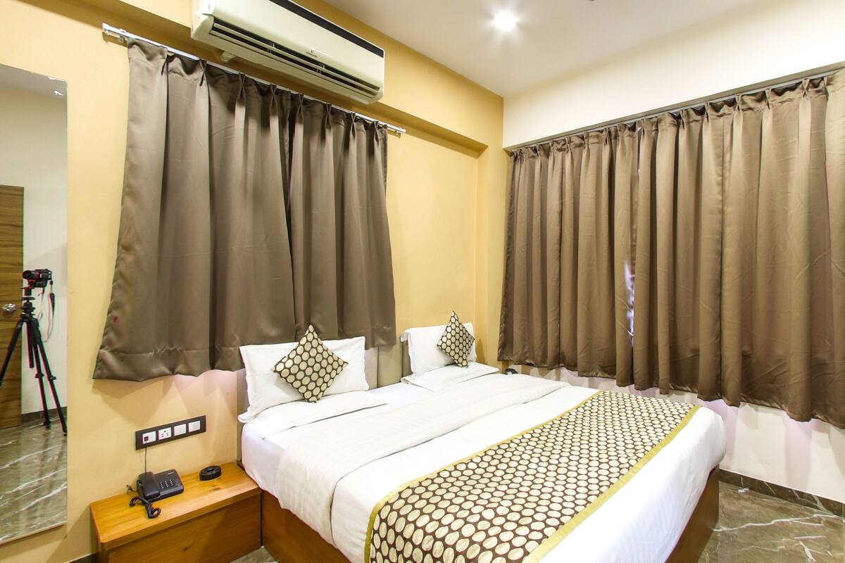 A hotel room with a bed, curtains, an air conditioning unit, a bedside table with a phone, and a wooden door.