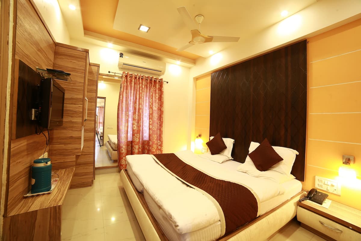 A hotel room with a large bed, a TV, a bedside table, a nightstand, and an air conditioning unit.