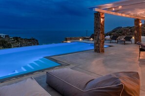 A poolside area with stone pillars, lounge chairs, and a view of the sea at dusk. - Villa Artemis By Mykonos Luxury (Mykonos)