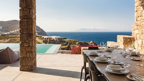 A stone pillar, a dining table set with plates and glasses, and a view of the sea and hills. - Villa Artemis By Mykonos Luxury (Mykonos)