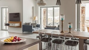 A modern living room with a wooden dining table, clear chairs, a TV mounted on the wall, and a stone wall visible through the window. - Villa Artemis By Mykonos Luxury (Mykonos)