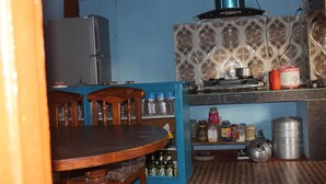 A kitchen with a refrigerator, stove, and various kitchen appliances. There is a dining table with chairs and a blue cabinet with bottles. - Buddha Shanti Lodge Backpackers paradise (Dhulikhel)