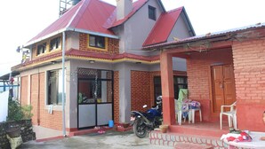 A two-story house with a red roof, a brick exterior, and a small porch with a table and chairs. - Buddha Shanti Lodge Backpackers paradise (Dhulikhel)