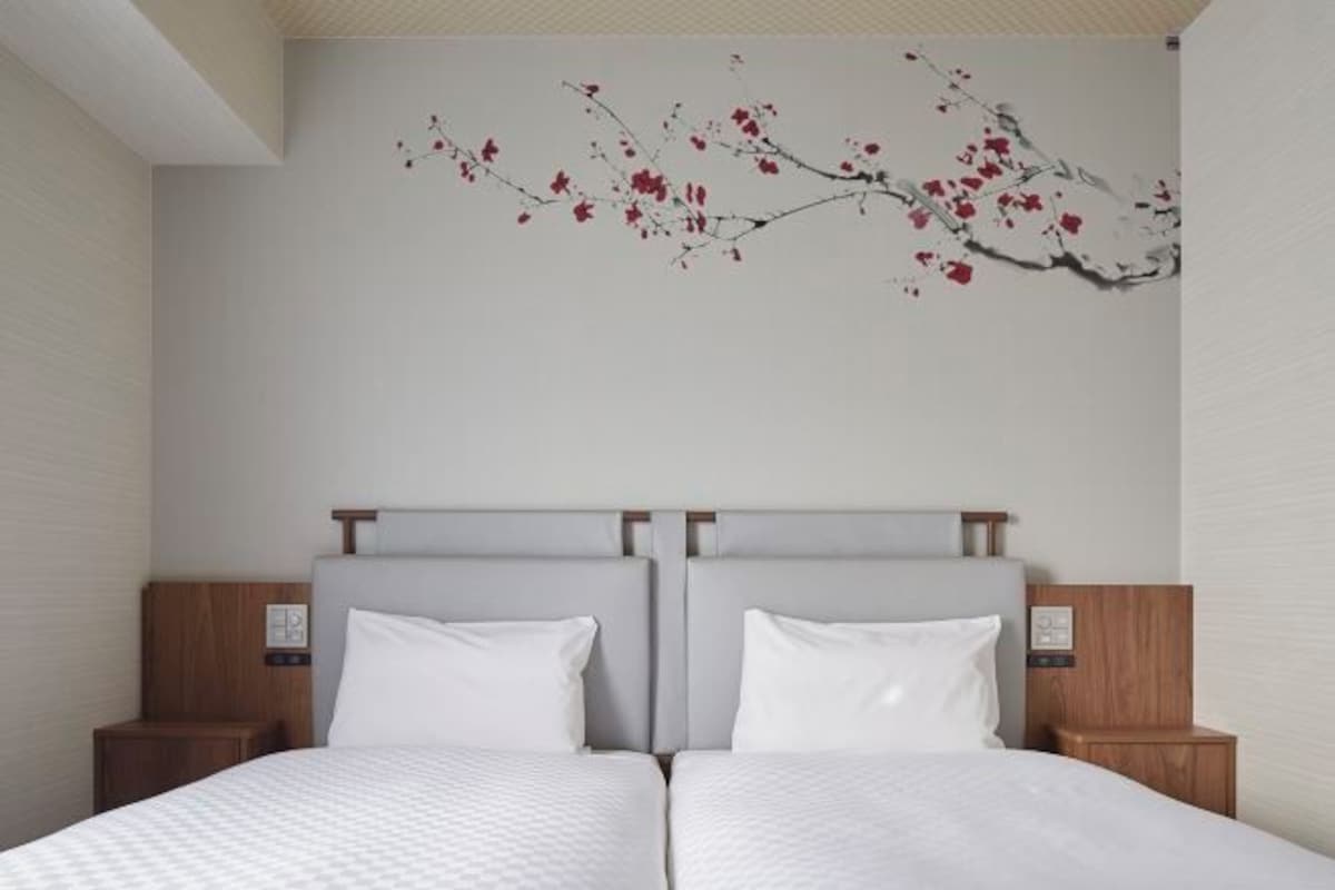 Una cama doble con cabecera gris, dos mesitas de noche y un cuadro colgado en la pared con una rama y flores rojas.
