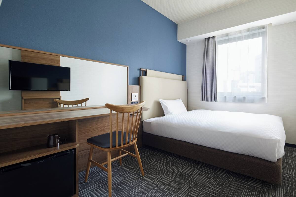 Una habitación de hotel con cama, escritorio, silla, televisor y ventana con cortinas.