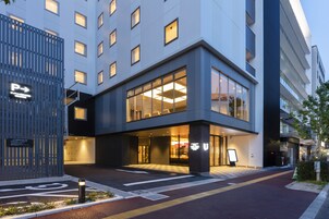 Una entrada de hotel moderna con un amplio ventanal de vidrio, un letrero de estacionamiento y un camino despejado que lleva a la entrada.