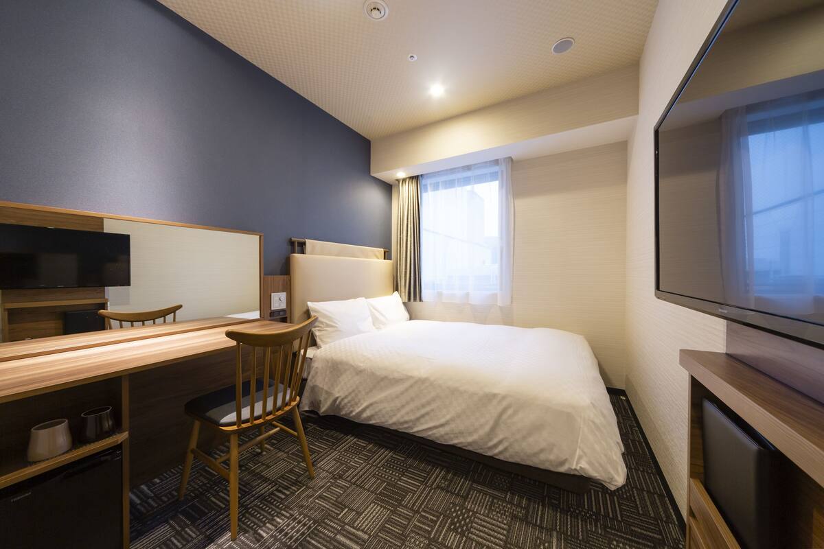 Habitación de hotel con cama, escritorio, silla, televisor y ventana con cortinas.