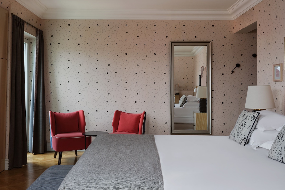 Una habitación de hotel con cama, una silla roja, una mesita, un espejo y una lámpara.