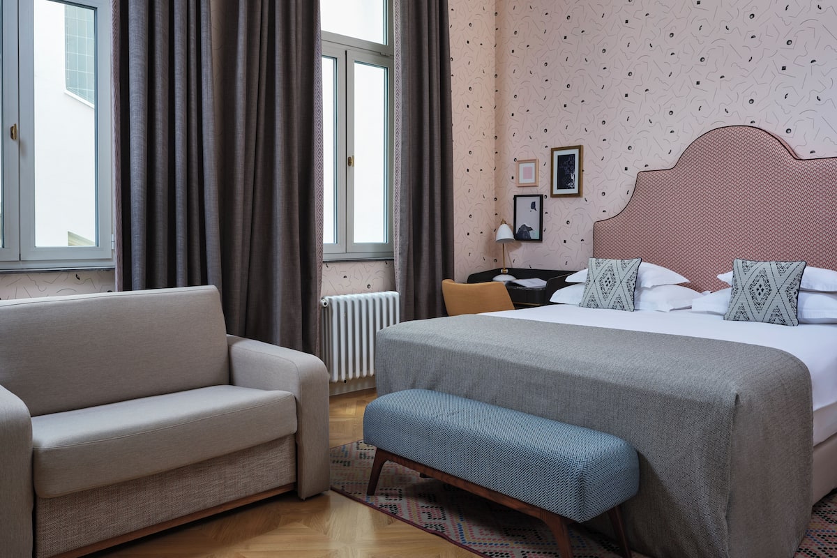Ein Hotelzimmer mit Bett, Sofa, Bank, Schreibtisch und einem Fenster mit Vorhängen.