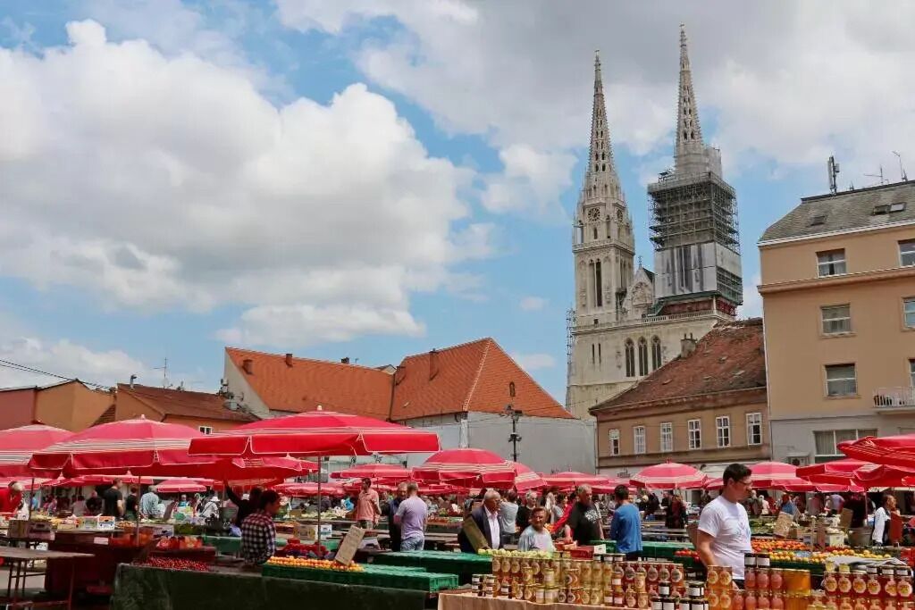 A bustling outdoor market with red umbrellas, surrounded by historic buildings and a prominent church tower.