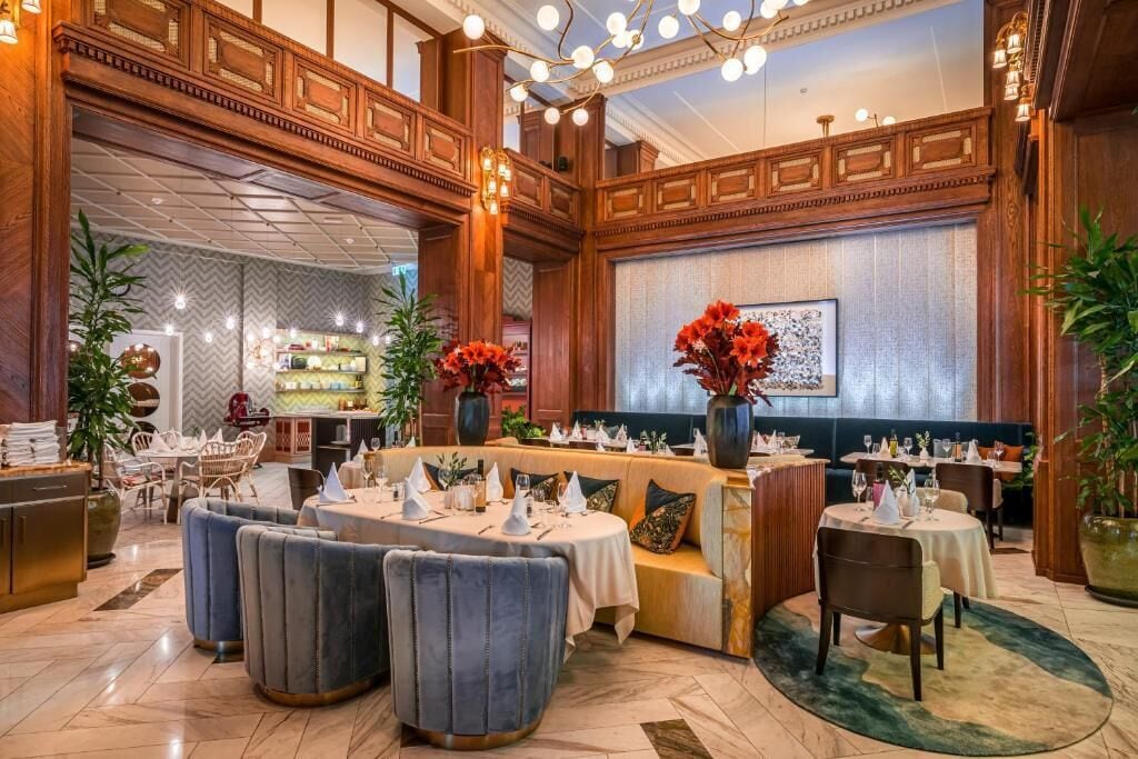 A spacious dining area with high wooden ceilings, multiple seating arrangements including round tables and sofas, and decorative lighting fixtures.