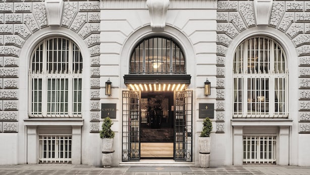 Une grande entrée avec une arche centrale, flanquée de deux larges fenêtres en arc de cercle, et un escalier menant à une porte surmontée d’un portail en métal.