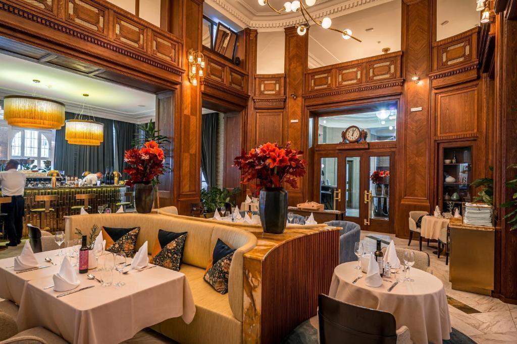 A luxurious hotel lobby with high ceilings, ornate wood paneling, and elegant chandeliers. There are multiple dining areas with round tables set for guests.