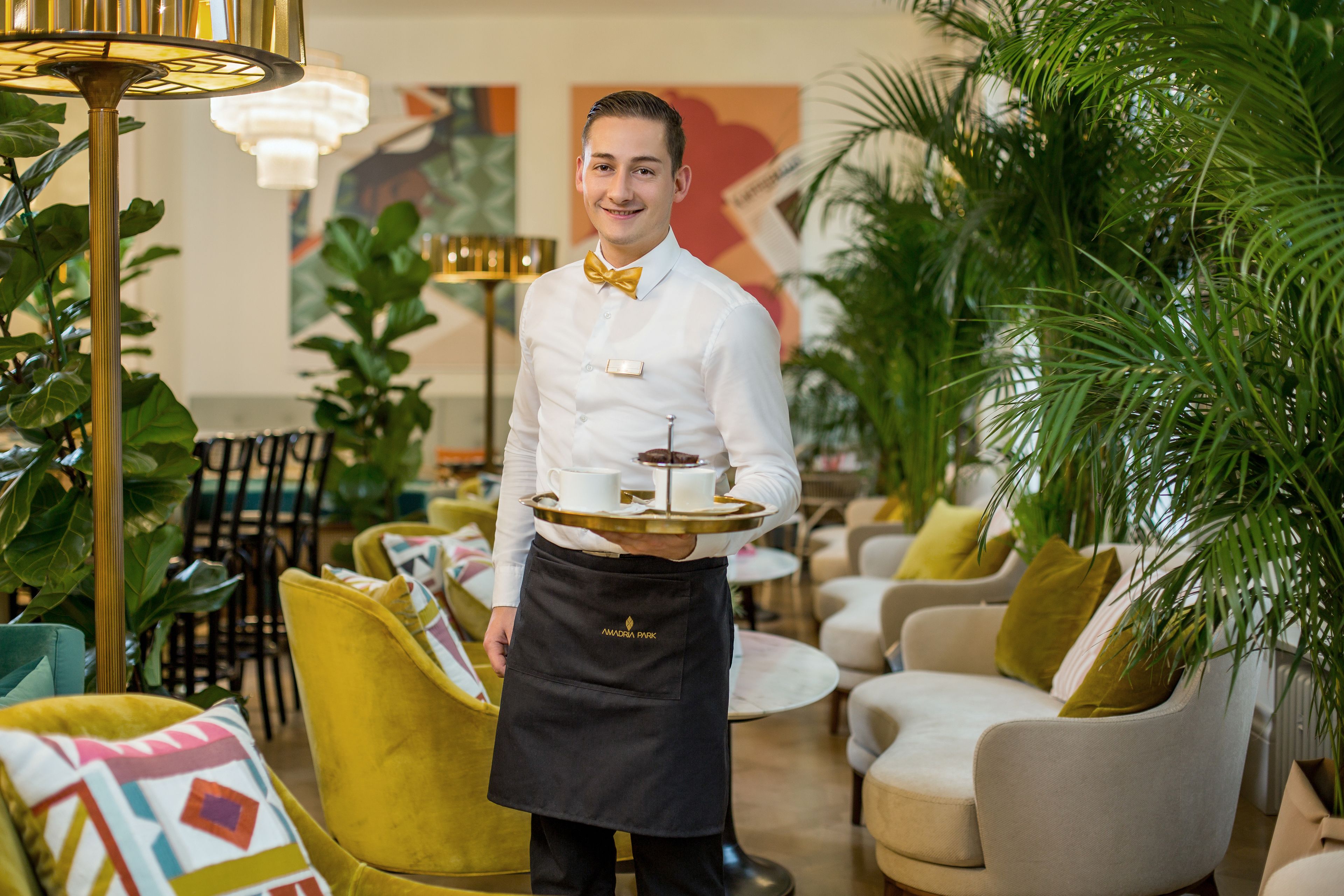 A waiter in a white shirt and black apron holding a tray with a teapot and cups in a well-lit indoor setting with green plants and comfortable seating.