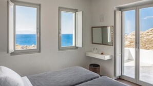 Une chambre avec deux lits, une fenêtre donnant sur la mer, une petite étagère blanche avec un miroir et une poubelle.
