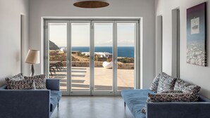 Un salon moderne doté d’une grande porte-fenêtre coulissante donnant sur une terrasse avec vue sur la mer et les bâtiments. On y trouve deux canapés avec des coussins à motifs et un lampadaire.