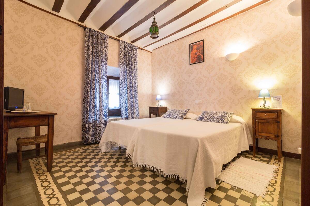 A bedroom with a checkered floor, a bed with white bedding, a wooden desk, a chair, a lamp, and a patterned curtain.