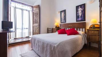 Un dormitorio con una cama grande, dos almohadillas rojas, un cabecero de madera, una mesita de noche con lámpara, un televisor sobre un mueble y una ventana con cortinas.
