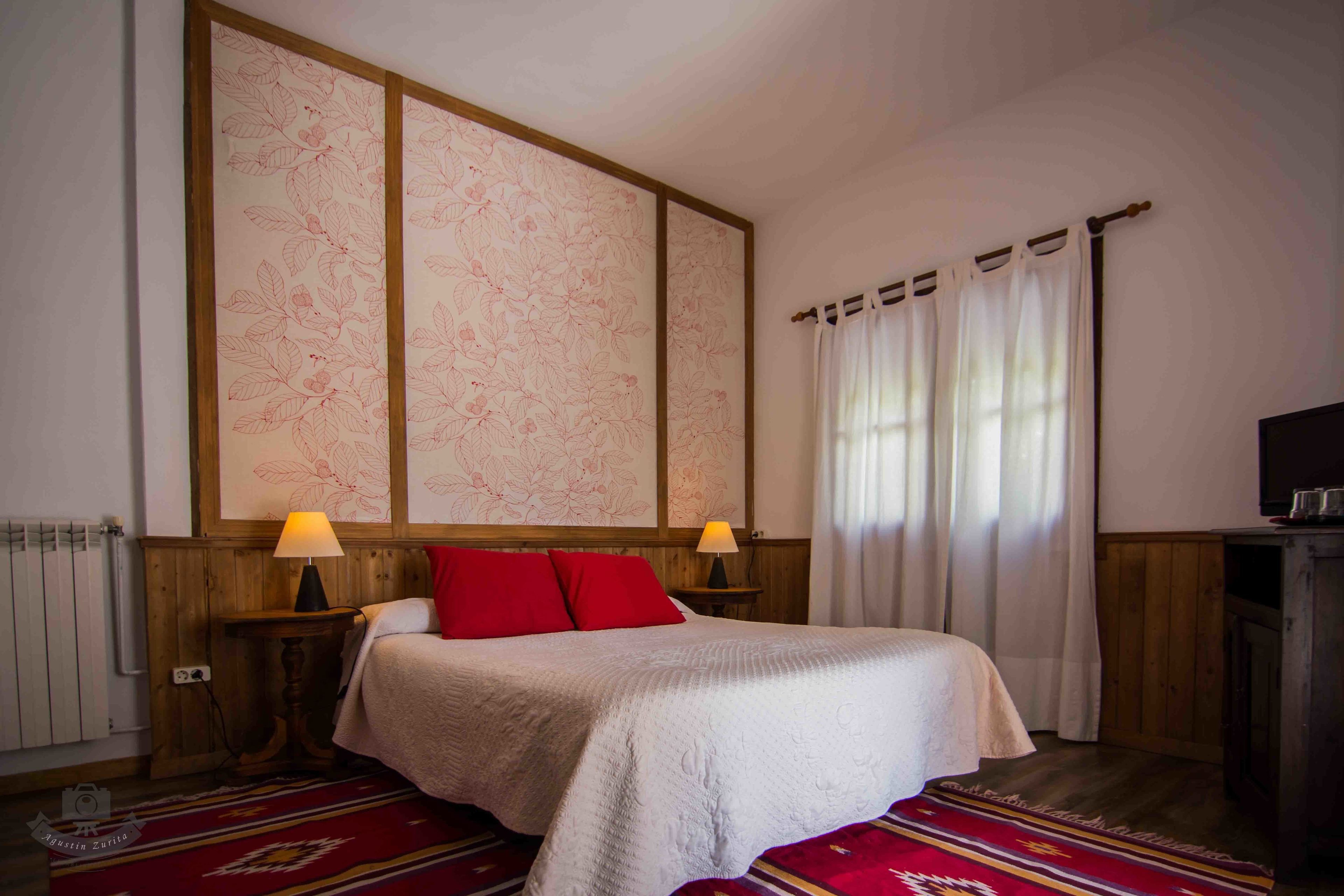 A bedroom with a bed, two bedside lamps, a radiator, a wooden headboard, a window with curtains, and a television on a wooden cabinet.