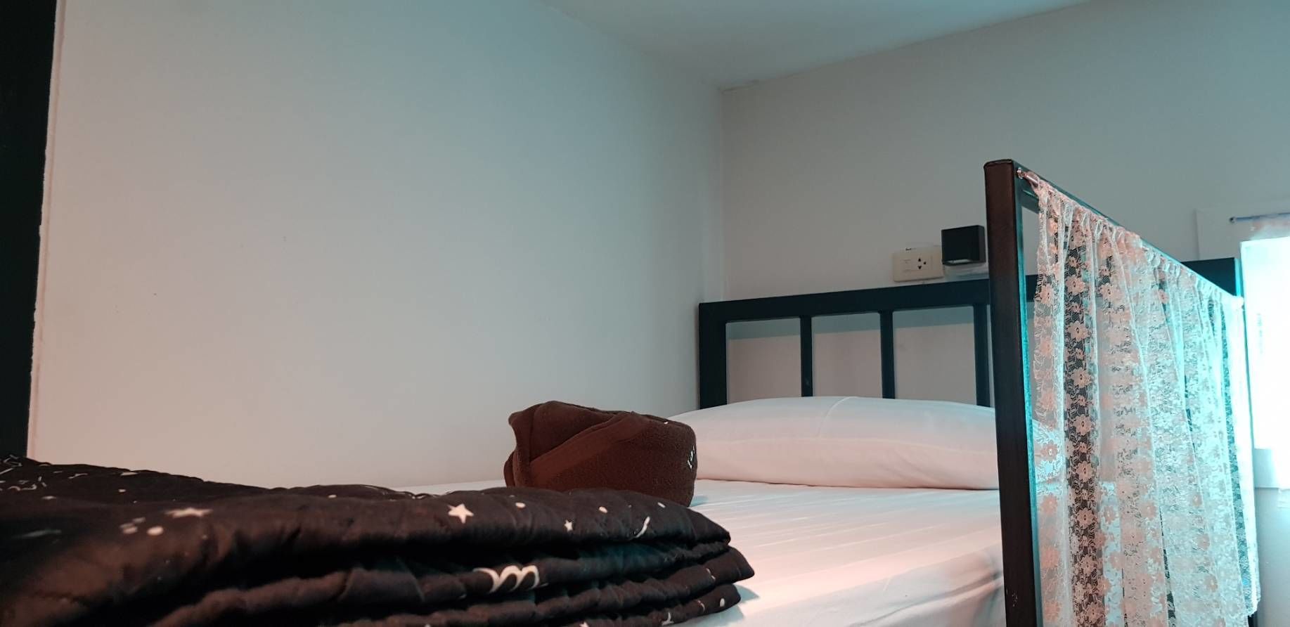 Una cama individual con un edredón oscuro y una almohada marrón, un cabecero de madera con motivos florales, y una pared blanca lisa al fondo.