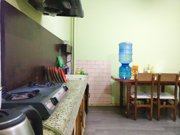 Una cocina con estufa a gas, lavamanos y dispensador de agua. Se observa una mesa de comedor con sillas y una pared de ladrillo al fondo.
