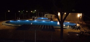 A swimming pool at night with surrounding lights and a tree in the foreground.