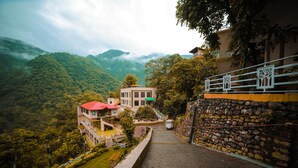 A hillside hotel with a red-roofed building, a stone retaining wall, and a view of lush green mountains. - Veda5 Ayurveda And Yoga Retreat (Yamkeshwar)