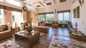 A spacious living area with multiple seating options, large windows offering views of greenery, and a mix of natural and artificial lighting. - Veda5 Ayurveda And Yoga Retreat (Yamkeshwar)
