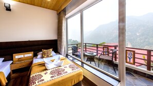 A hotel room with a large window offering a mountain view, a bed with a decorative headboard, a small table, and a chair on the balcony. - Veda5 Ayurveda And Yoga Retreat (Yamkeshwar)