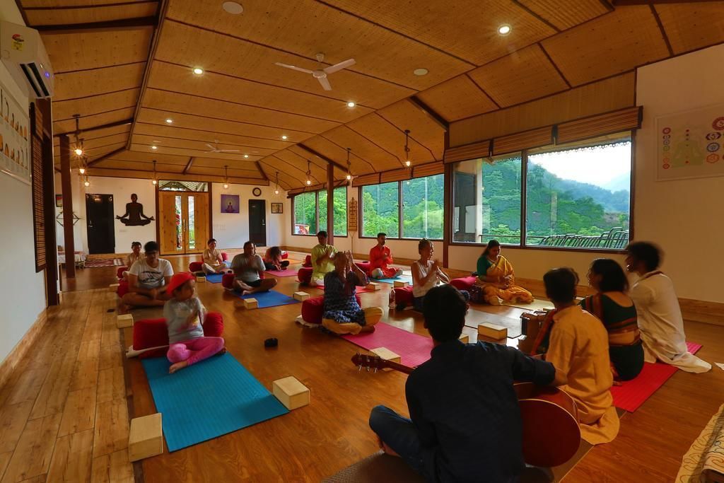 A yoga class in a room with wooden floors and large windows, participants seated on mats.